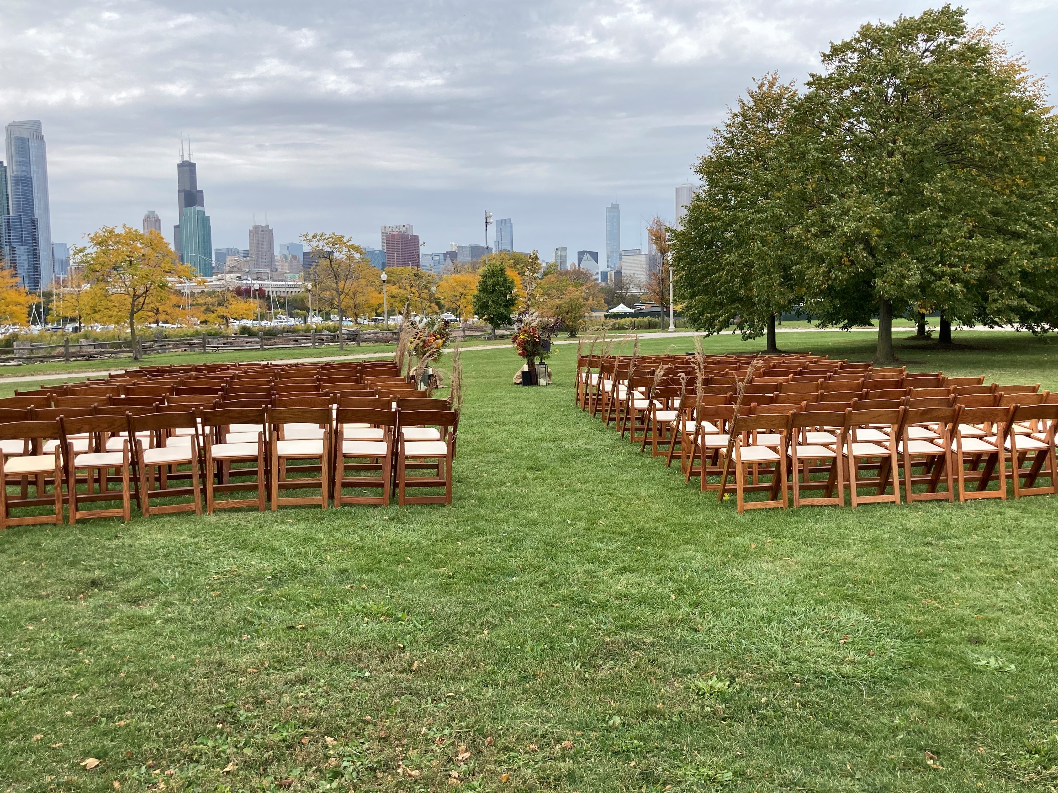 Outdoor wedding venue with rows of empty wooden chairs on a grassy lawn, Chicago skyline in background.